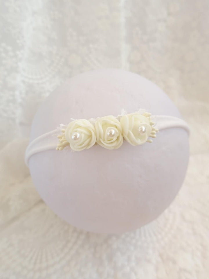 A white headband adorned with three white roses is displayed against a white lace background.