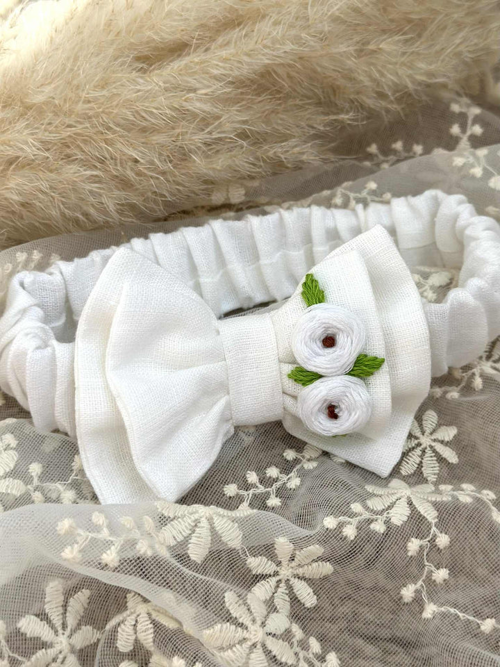 A white headband with a bow and two white flowers is displayed on a white lace background.