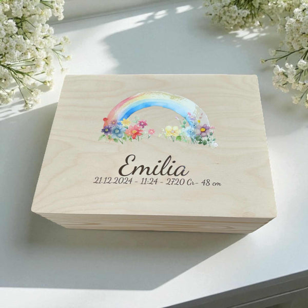 A wooden box with a colorful rainbow graphic and the name "Emilia" printed on it, placed on a white surface with white flowers in the background.