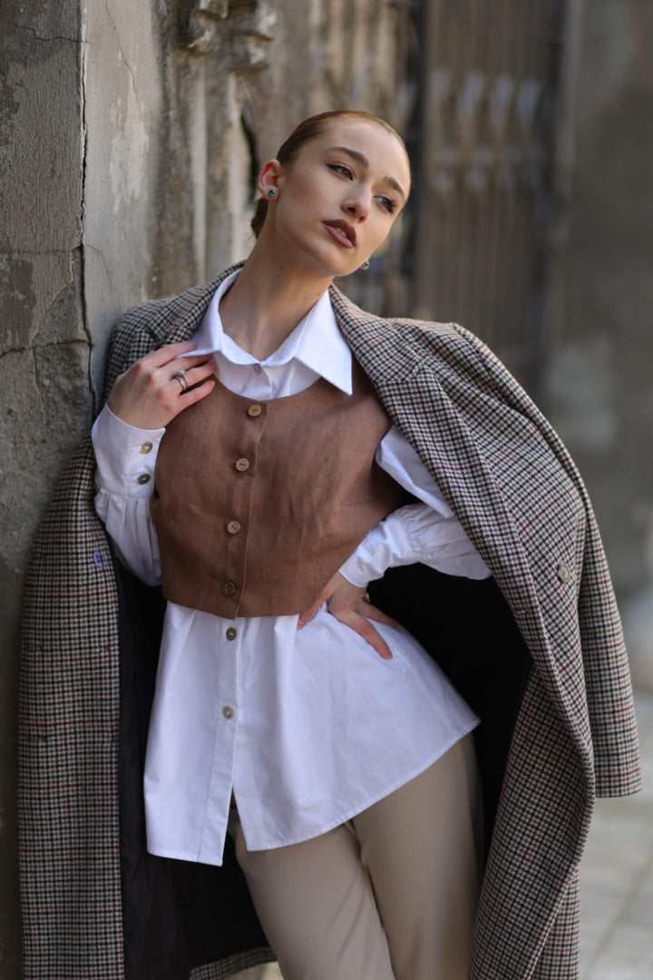 A woman in a brown vest and white blouse stands against a stone wall, leaning against it with her hands on her hips.