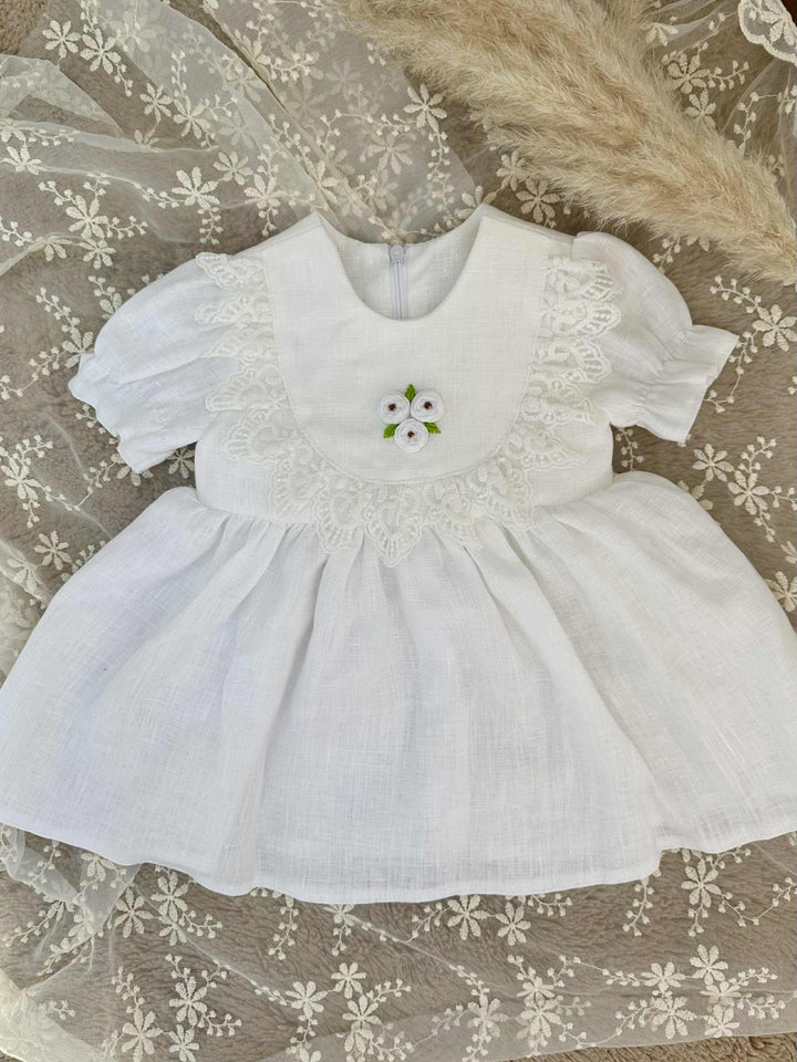 A white lace dress with a floral embroidered bodice and a ruffled collar, placed on a white lace background.