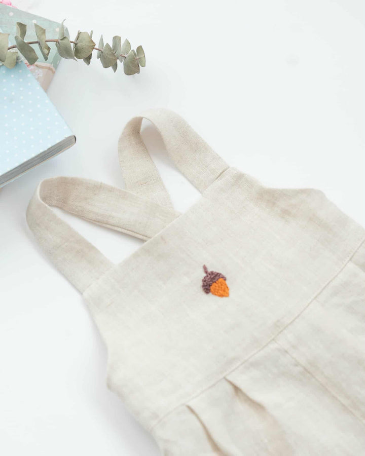 A beige dress with a small embroidered acorn detail is displayed on a white surface, accompanied by a small plant and a blue book.