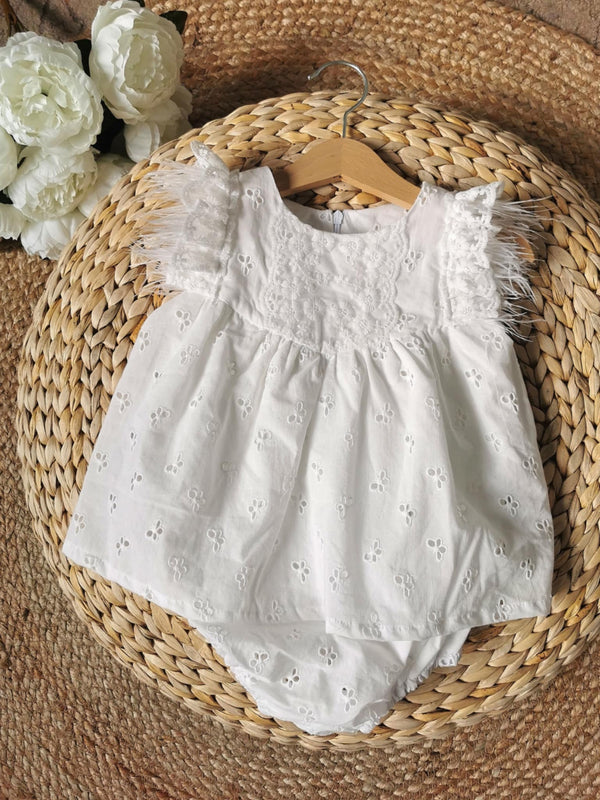 A white baby dress with lace details and feather trim is displayed on a wooden hanger, placed on a woven mat.