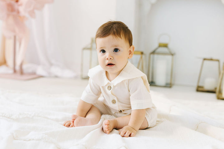 A baby boy in a white outfit sits on a white blanket, looking directly at the camera with a curious expression.