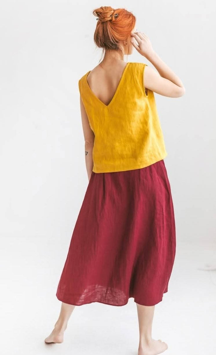 A woman with red hair wearing a yellow sleeveless top and a maroon skirt stands with her back to the camera, her hand on her head.