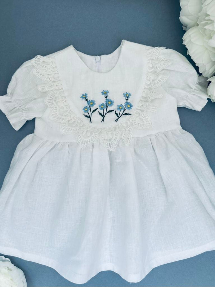 A white dress with blue flowers embroidered on the front, displayed on a blue background.