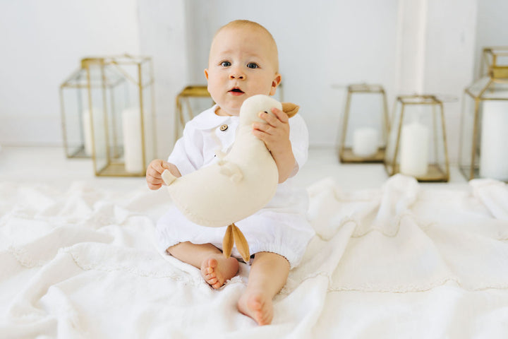 A baby in a white outfit is sitting on a white blanket, holding a stuffed animal.