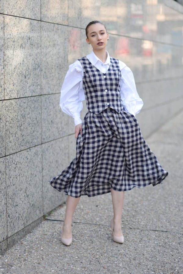 A woman wearing a white blouse and a blue and white checkered dress stands in front of a stone wall, posing for the camera.