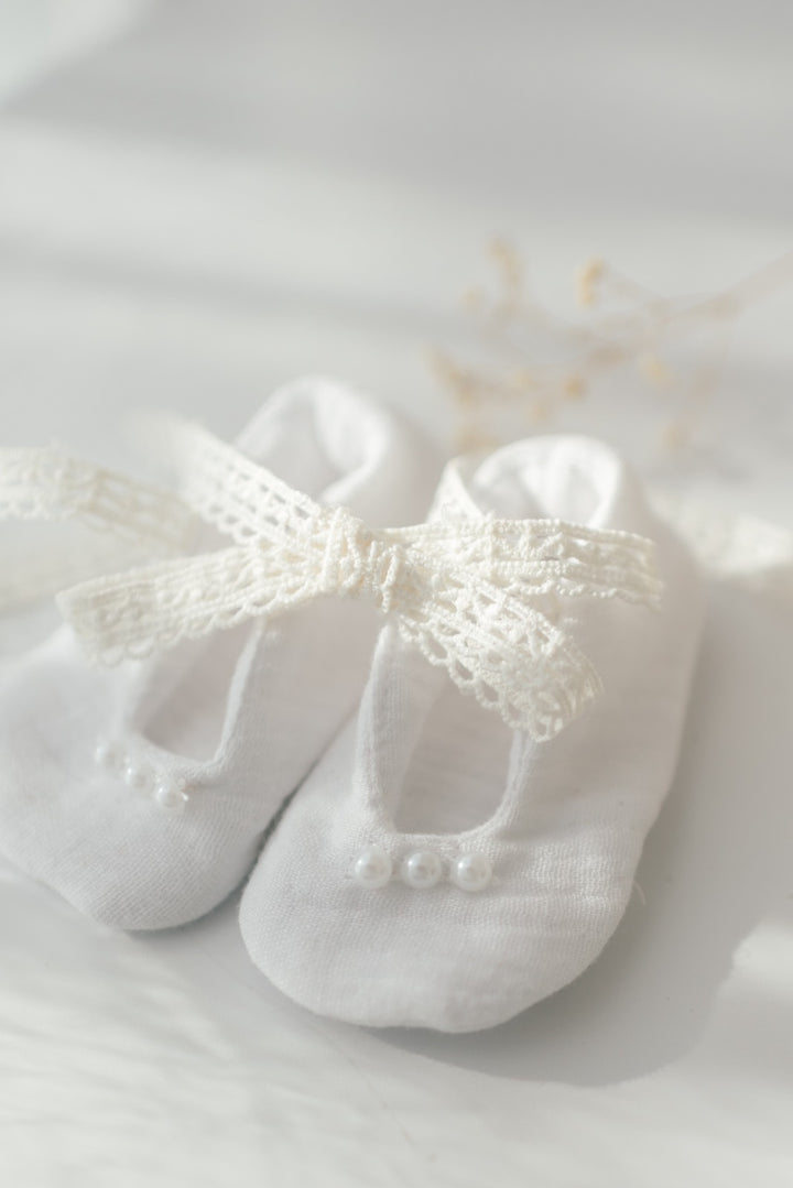A pair of white baby shoes with a delicate lace bow tied around the toe area, resting on a white surface.