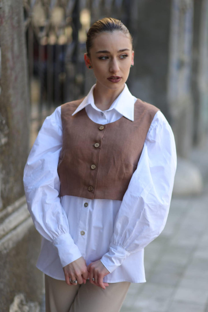 A woman wearing a brown vest, white shirt, and khaki pants stands in front of a stone wall, looking off to the side with a thoughtful expression.