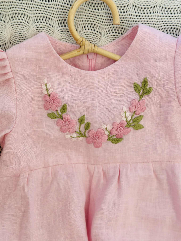A pink dress with a floral embroidered design on the front, hanging on a wooden hanger.