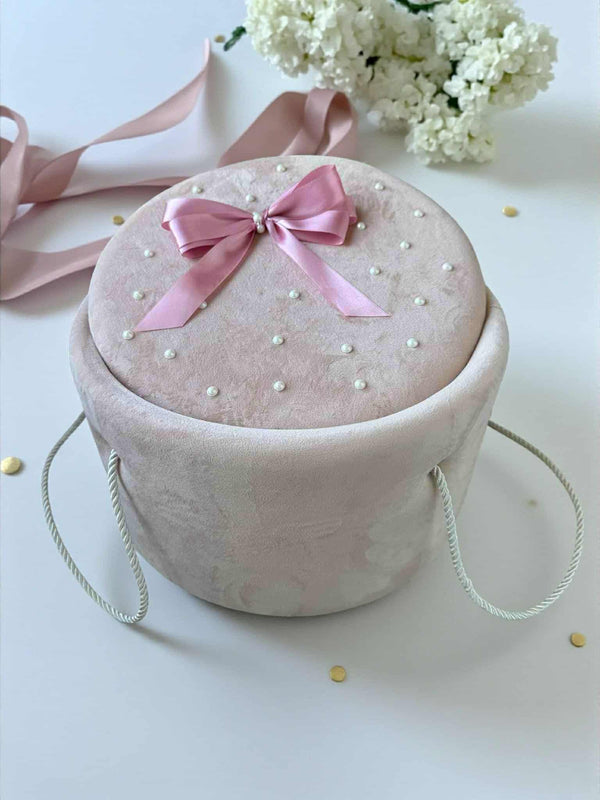 A pink velvet box with a pink ribbon and white pearls sits on a white surface, surrounded by scattered gold dots.