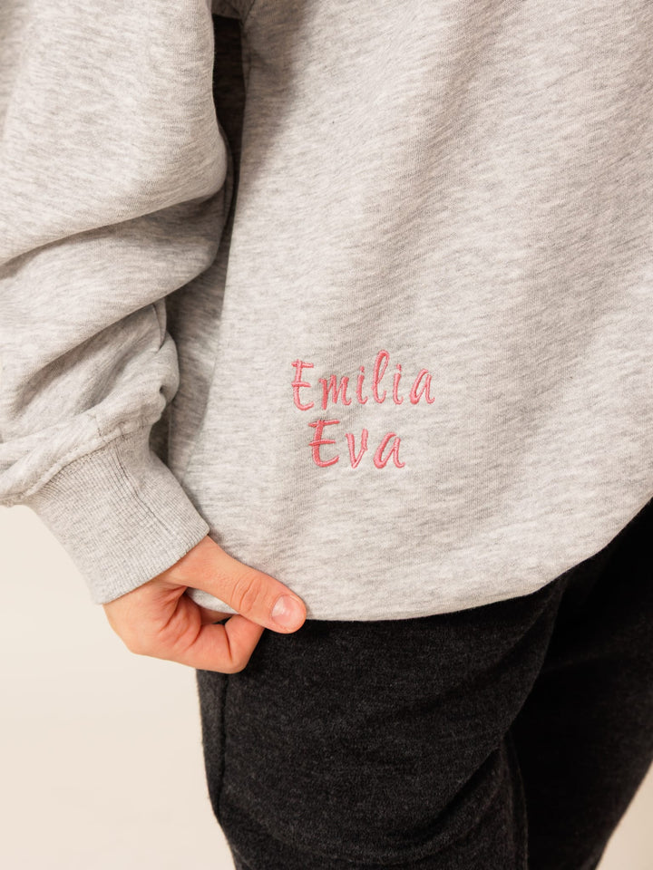 A person wearing a grey sweatshirt with the name 'Emilia' embroidered on it in pink.