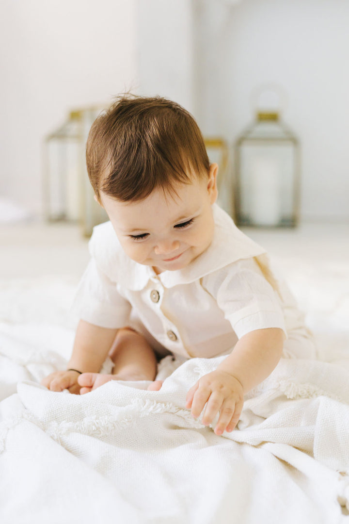 A smiling baby in a white outfit sits on a white blanket, reaching out to touch the blanket.