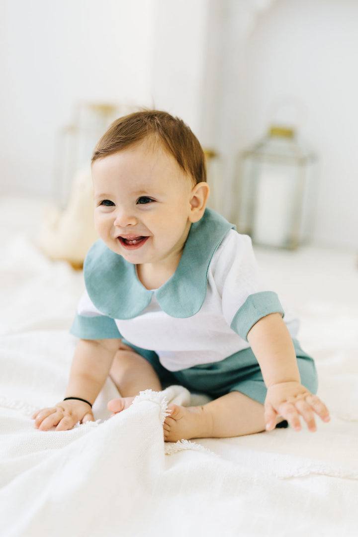 A smiling baby boy wearing a white and blue outfit sits on a white blanket, looking directly at the camera with a joyful expression.