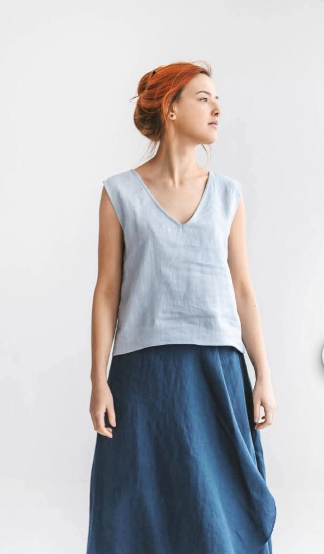 A woman with red hair wearing a gray sleeveless top and a blue skirt stands against a white background.