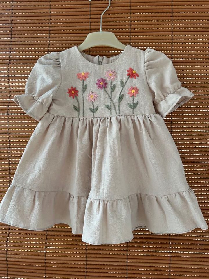 A beige dress with ruffled sleeves and a floral embroidered design on the front, hanging on a wooden hanger.