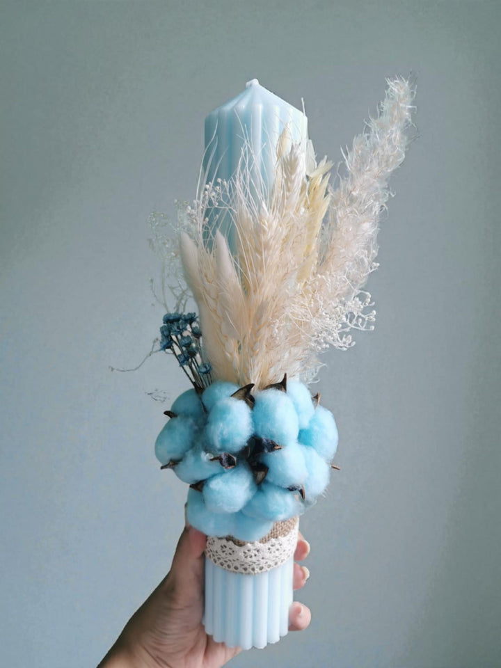 A hand is holding a blue candle holder with a white candle and various dried flowers and pom poms.