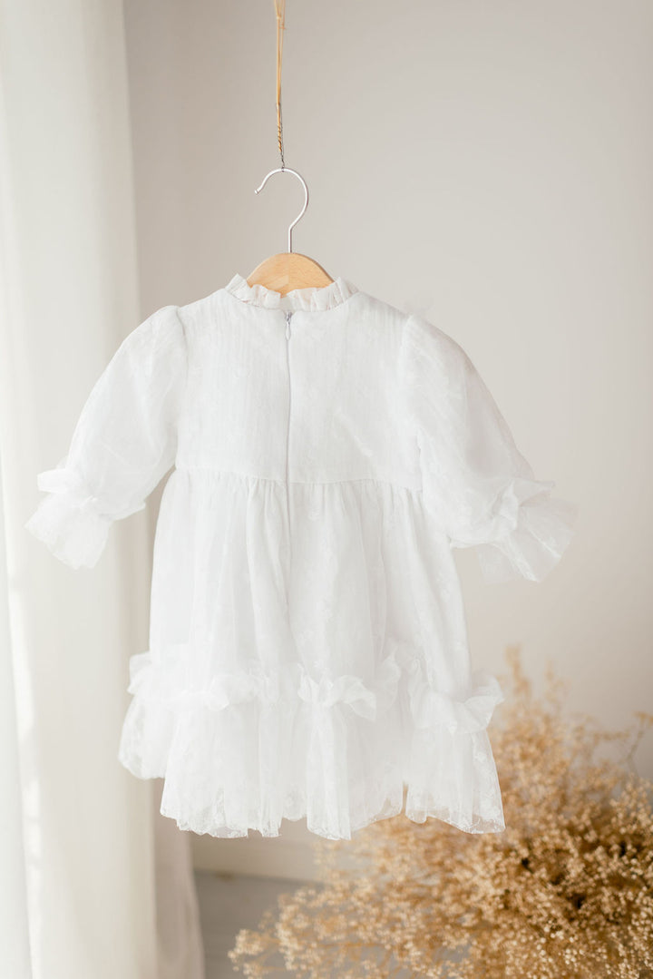 A white, ruffled dress is hanging on a wooden hanger against a white wall.