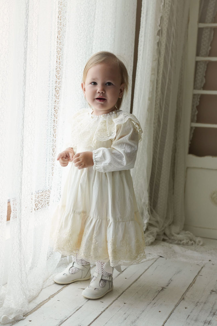 A young girl in a white lace dress stands in front of a window, gazing at the camera with a thoughtful expression.