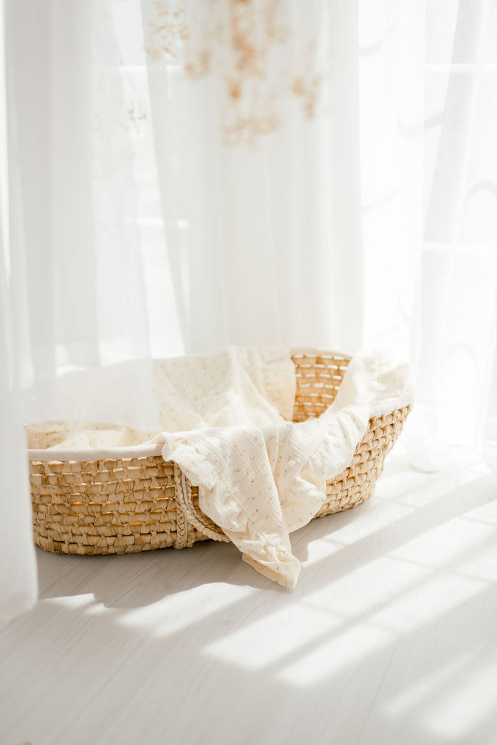A woven basket filled with a soft, white blanket sits on a wooden floor, with a window and white curtains in the background.