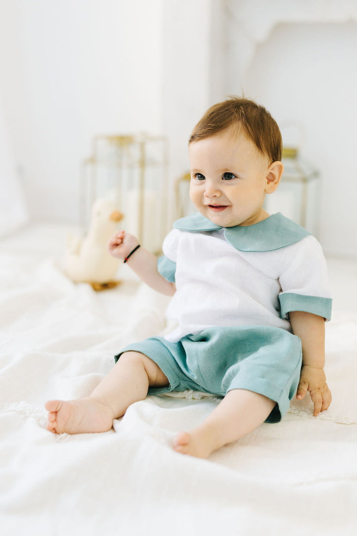 A smiling baby boy in a white and green outfit sits on a white blanket, with a stuffed animal in the background.
