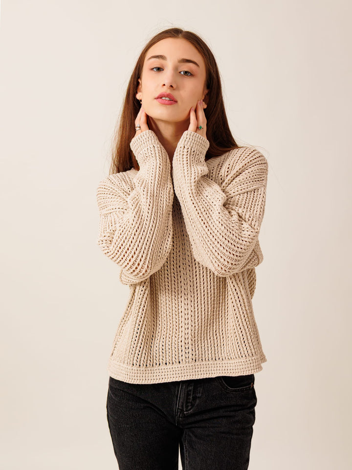 A young woman with long brown hair stands against a plain background, wearing a beige sweater and black pants. She has her hands on her face, looking directly at the camera with a thoughtful expression.