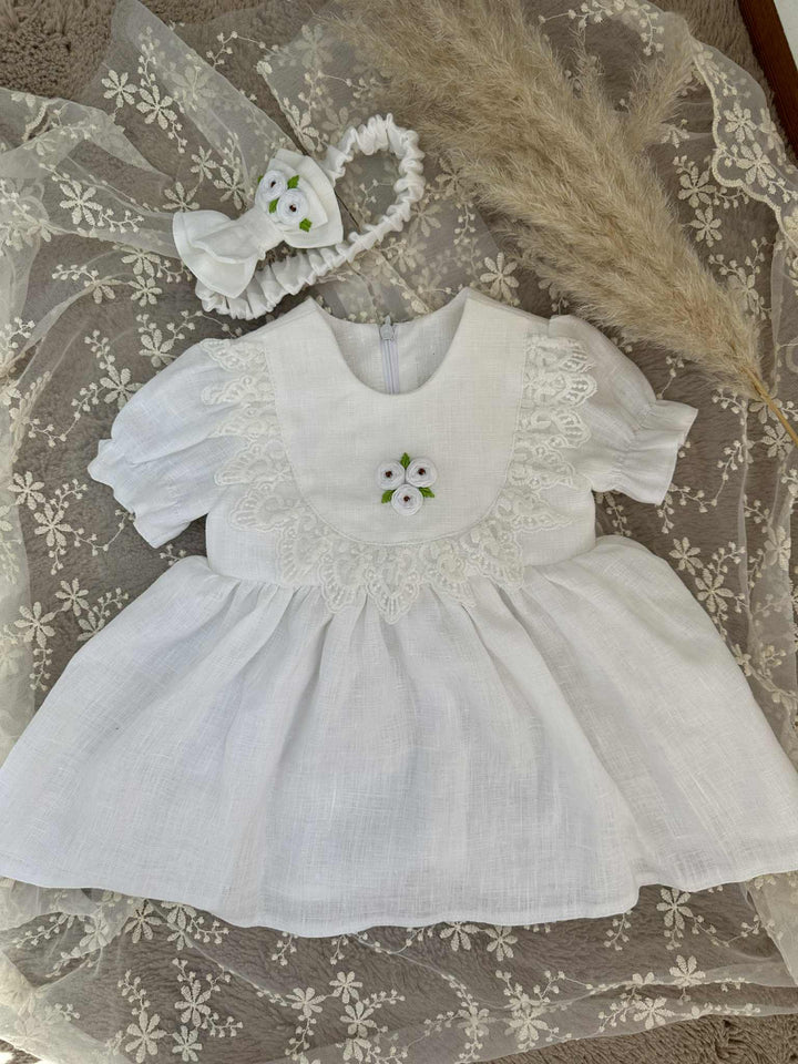A white lace dress with a floral embroidered bodice and a white headband is displayed on a white lace background.