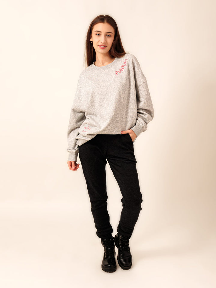 A young woman wearing a grey sweatshirt with a red logo, black pants, and black boots stands against a white background.