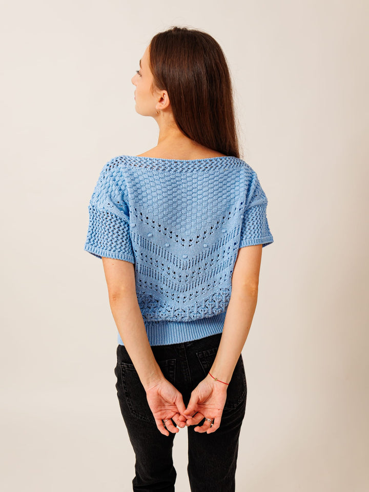 A woman with long brown hair is standing with her hands behind her back, wearing a blue knitted sweater.