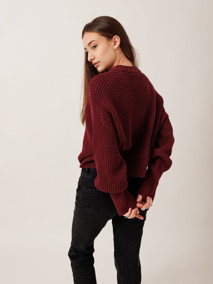 A young woman wearing a burgundy sweater and black jeans stands with her hands behind her back, looking away from the camera.