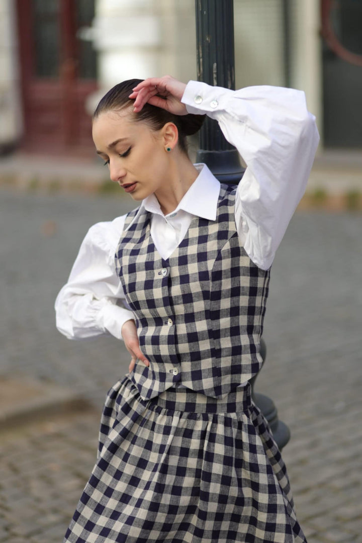 A woman in a white and black checkered dress stands on a cobblestone street, her hand resting on her head.