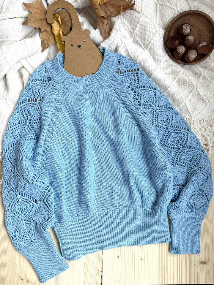 A light blue knitted sweater with intricate lace patterns is displayed on a wooden surface, accompanied by a wooden hanger and a bowl of small round objects.