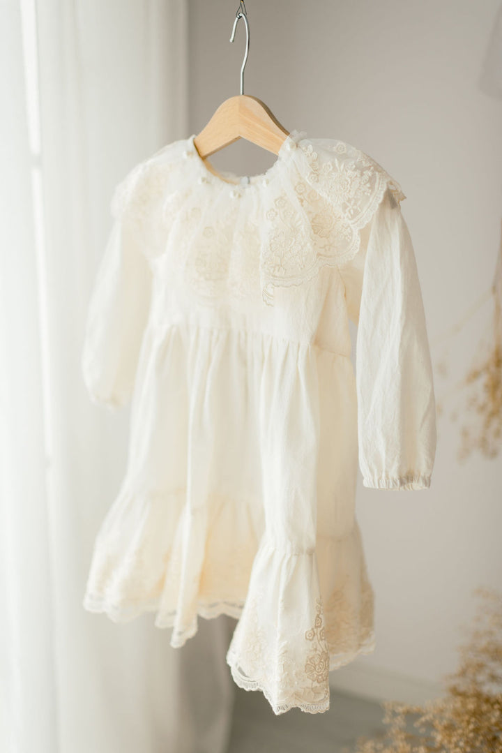 A white lace dress with ruffled sleeves is hanging on a wooden hanger against a white wall.