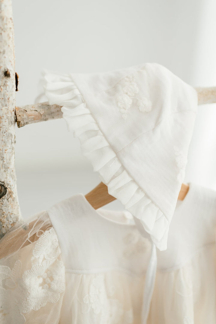 A white lace dress with a ruffled collar and a white bonnet draped over a wooden hanger.