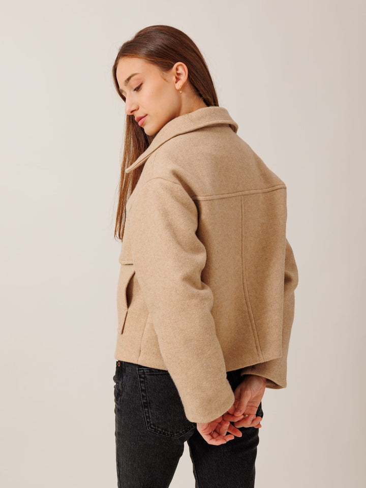 A woman with long brown hair wearing a beige coat stands with her hands behind her back, looking down.