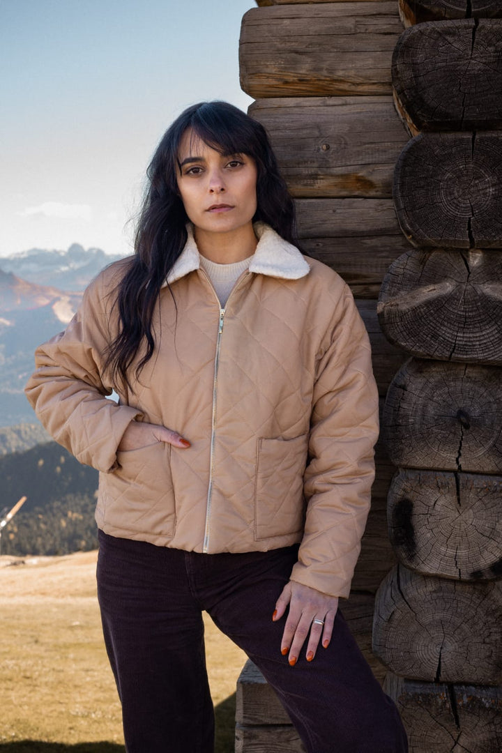 A woman in a tan jacket stands in front of a wooden log cabin, with a mountainous landscape visible in the background.