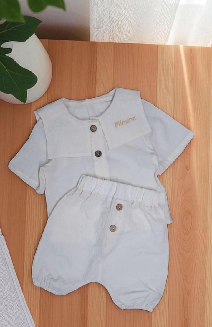 A white outfit consisting of a shirt and shorts is laid out on a wooden surface, with a potted plant visible in the background.