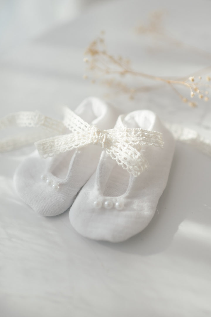 A pair of white baby shoes with a delicate lace bow sits on a white surface, with delicate white flowers in the background.