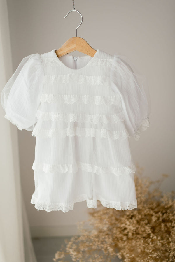 A white, ruffled dress with lace detailing is hanging on a wooden hanger against a white wall.
