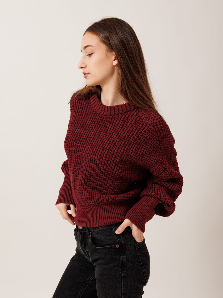 A woman wearing a burgundy sweater and black pants stands with her hands in her pockets, looking to her left.