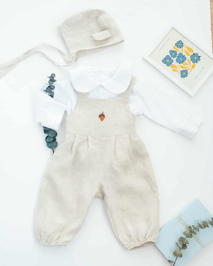 A beige outfit consisting of a white shirt, a beige hat, and a beige jumpsuit with a small orange flower on the front, along with a framed floral print, is laid out on a white surface.