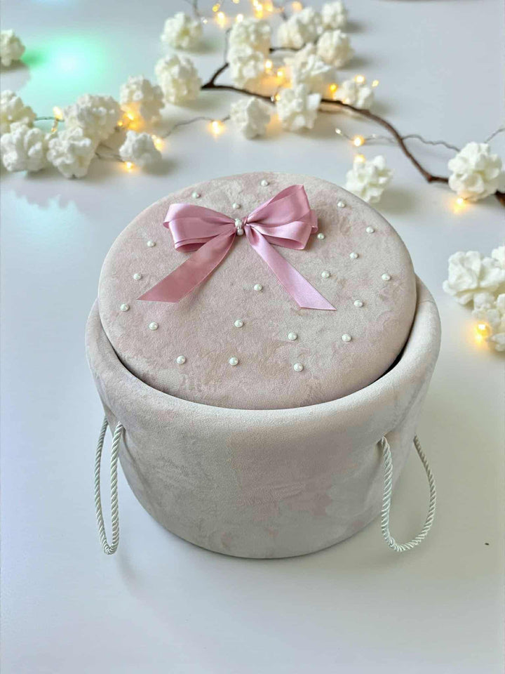 A pink velvet box with a bow sits on a white surface, surrounded by white flowers and twinkling lights.