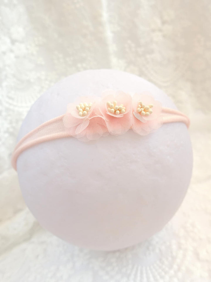A delicate pink flower headband with three petals and small white flowers is displayed against a white background.