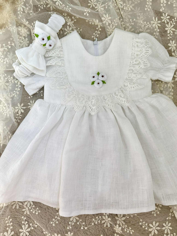 A white lace dress with a floral embroidered bodice and a white flower headband is displayed on a white lace background.