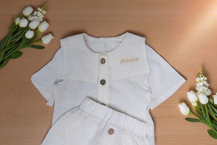 A white outfit with a name tag is displayed on a wooden surface, accompanied by a bouquet of white flowers.