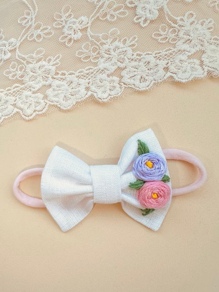 A white bow with pink and purple flowers is displayed on a beige background, accompanied by a delicate white lace fabric.