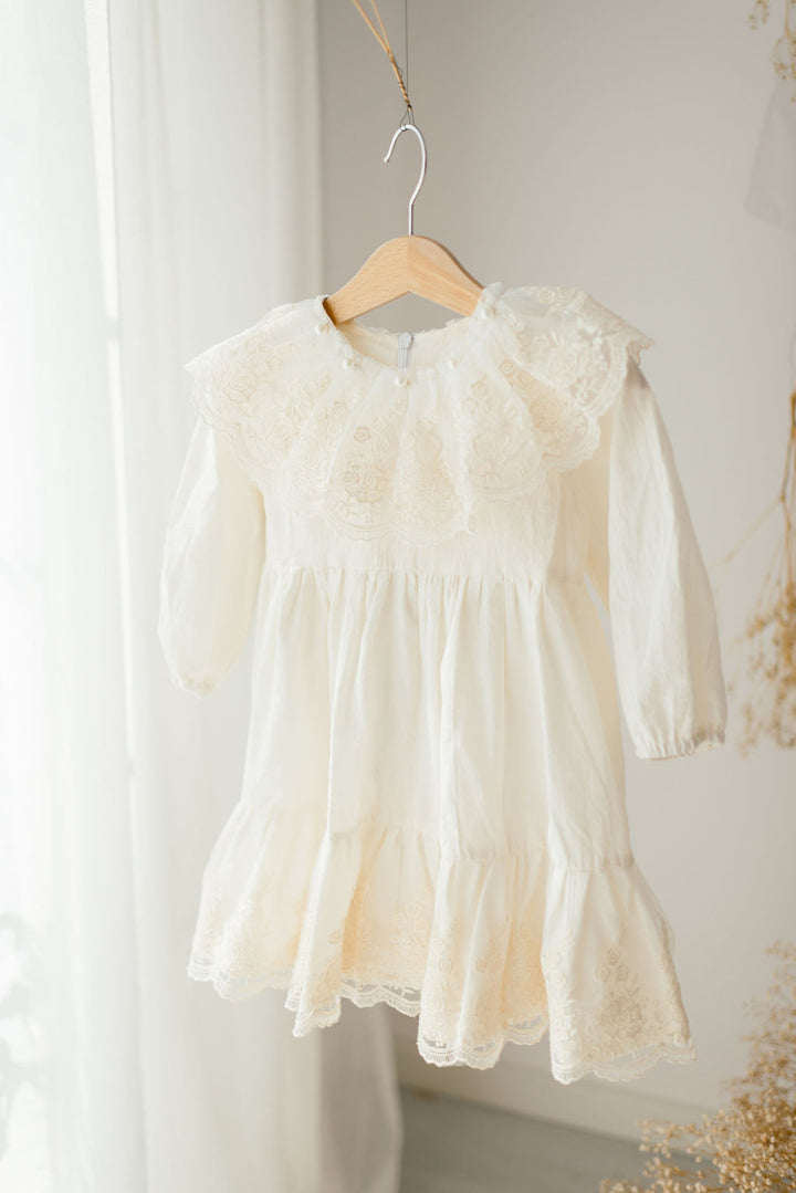 A white lace dress with ruffles is hanging on a wooden hanger against a white wall.