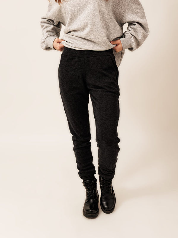 A young woman wearing a grey sweatshirt with a red logo, black pants, and black boots stands against a white background.