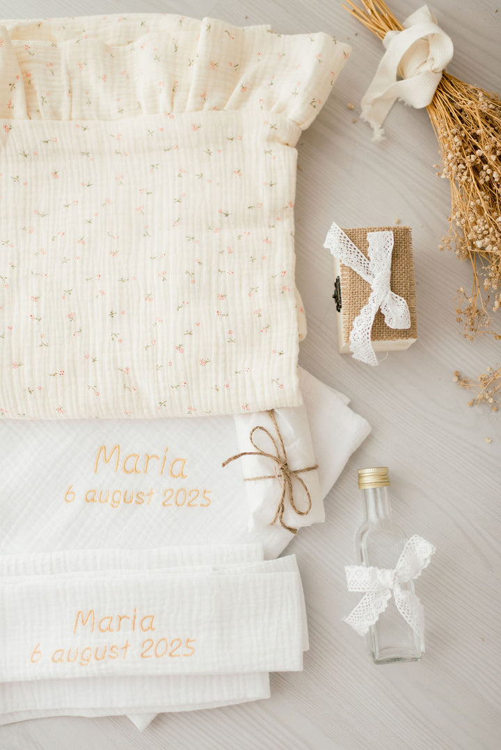 a collection of items related to a baby shower, including a white cloth with text, a small gift box, a white ribbon, and a small glass bottle with a white ribbon.